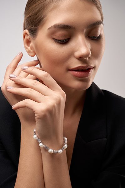 Crop of young beautiful smiling female model in formal suit presenting silver bracelet with white pearls. Portrait of woman with perfect makeup posing in studio, isolated on gray.